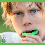 Young athlete putting in a green sports mouthguard, close-up of face with intense expression, wearing white sports gear — promoting custom mouthguards at Spring Hill Dental.